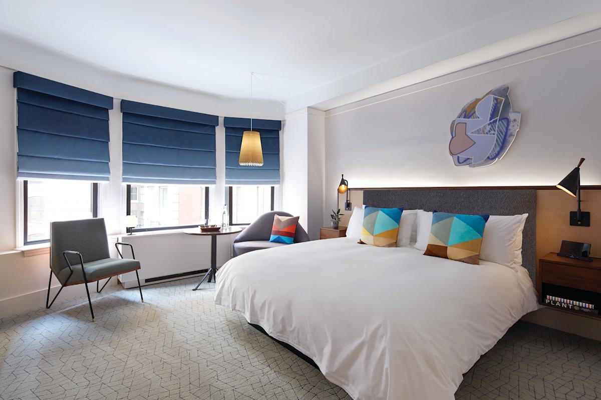 A modern hotel room features a large bed with geometric-patterned pillows, two chairs near bay windows with blue shades, a wall lamp, and contemporary artwork above the headboard.