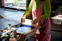 Person in a green shirt and pink apron using a screen printing squeegee on a table. Sunlight streams through a window, illuminating the workspace filled with printing equipment.