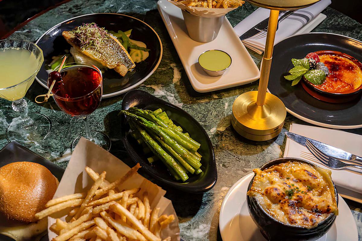 A table with a variety of dishes: grilled fish on greens, asparagus, French fries, a dinner roll, a bubbling cheesy casserole, crème brûlée, and two cocktails. The table has a green marble texture and a gold lamp.