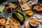A table with a variety of dishes: grilled fish on greens, asparagus, French fries, a dinner roll, a bubbling cheesy casserole, crème brûlée, and two cocktails. The table has a green marble texture and a gold lamp.