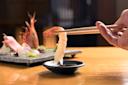 A hand uses chopsticks to dip a piece of shrimp sushi into a small dish of soy sauce, with a plate of assorted sashimi and sushi in the blurred background on a wooden table.