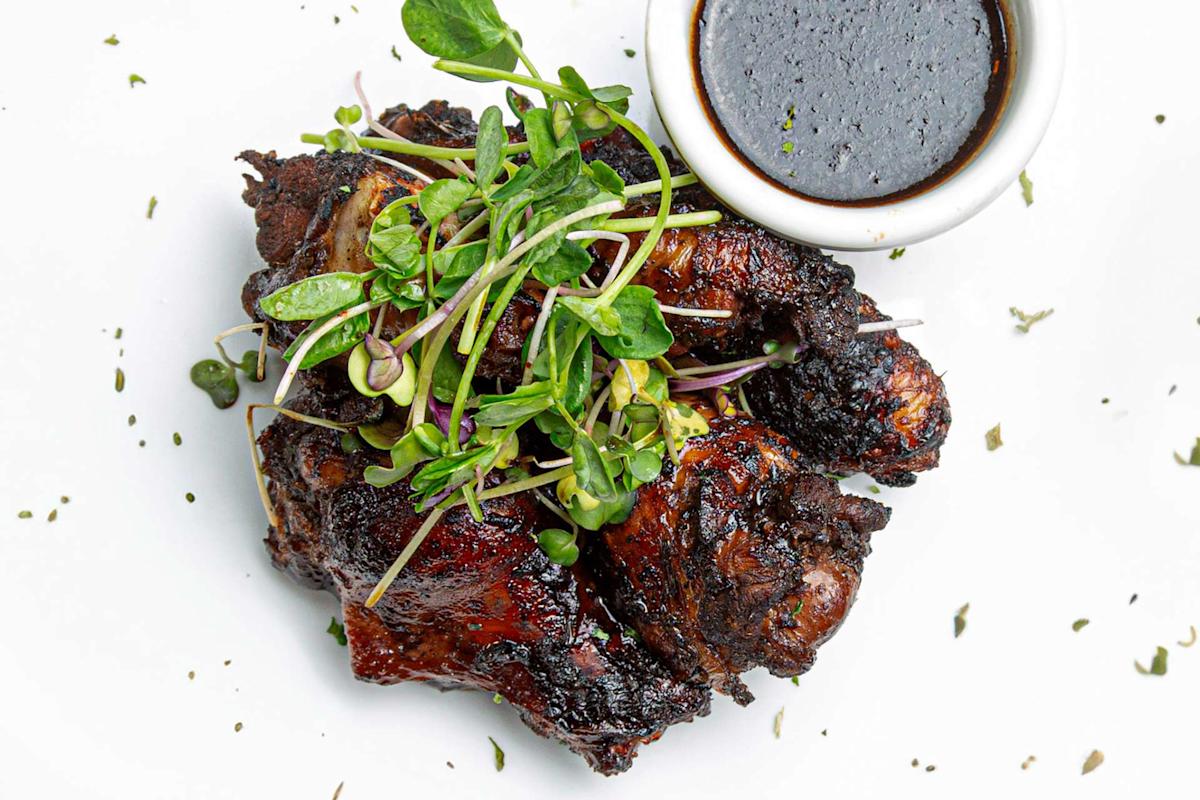 Charred pieces of grilled chicken topped with fresh microgreens, served on a white plate with a small bowl of dark dipping sauce on the side. Dried herbs are sprinkled around the dish.
