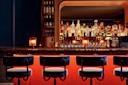 A dimly lit bar features several bottles of liquor arranged on shelves behind a marble counter. Four upholstered bar stools are lined up in front, and small lamps create a warm ambiance.