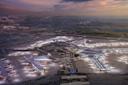Aerial view of a large, brightly lit airport at dusk, with multiple terminals, airplanes parked at gates, runways, and a cityscape in the background under a cloudy sky.
