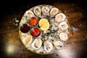 A round platter of raw oysters on ice, garnished with seaweed and lemon wedges, with three dipping sauces in metal cups, displayed on a dark wooden surface.