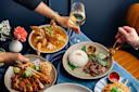 A table with a variety of Asian dishes, including a bowl of crispy fried crab, a plate of rice with beef and greens, and slices of roasted meat with noodles. Hands are reaching for the food and a glass of white wine.