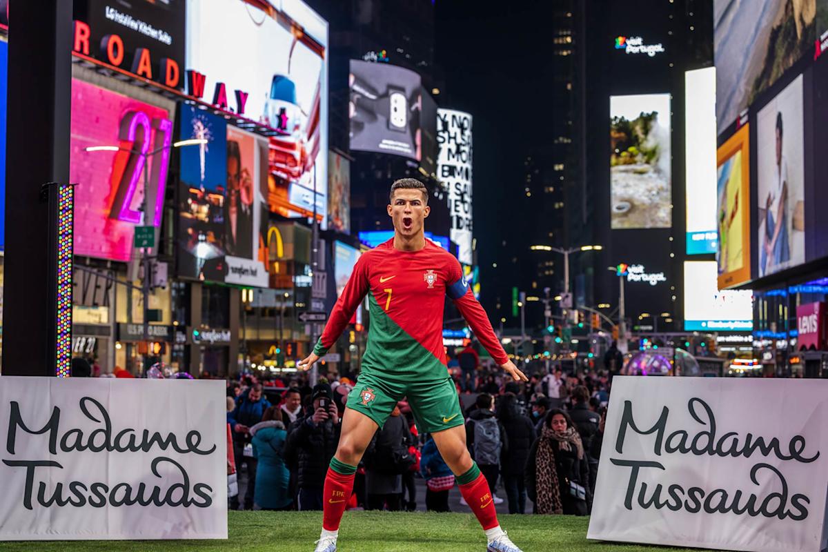 A wax figure of a Portuguese soccer player in a red and green uniform stands in Times Square at night, surrounded by crowds and bright billboards, with Madame Tussauds signs on both sides.