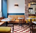 A cozy restaurant interior with tan leather banquette seating, round marble tables, wooden chairs, red and white checkered floor tiles, exposed brick wall, and a modern wall sconce.