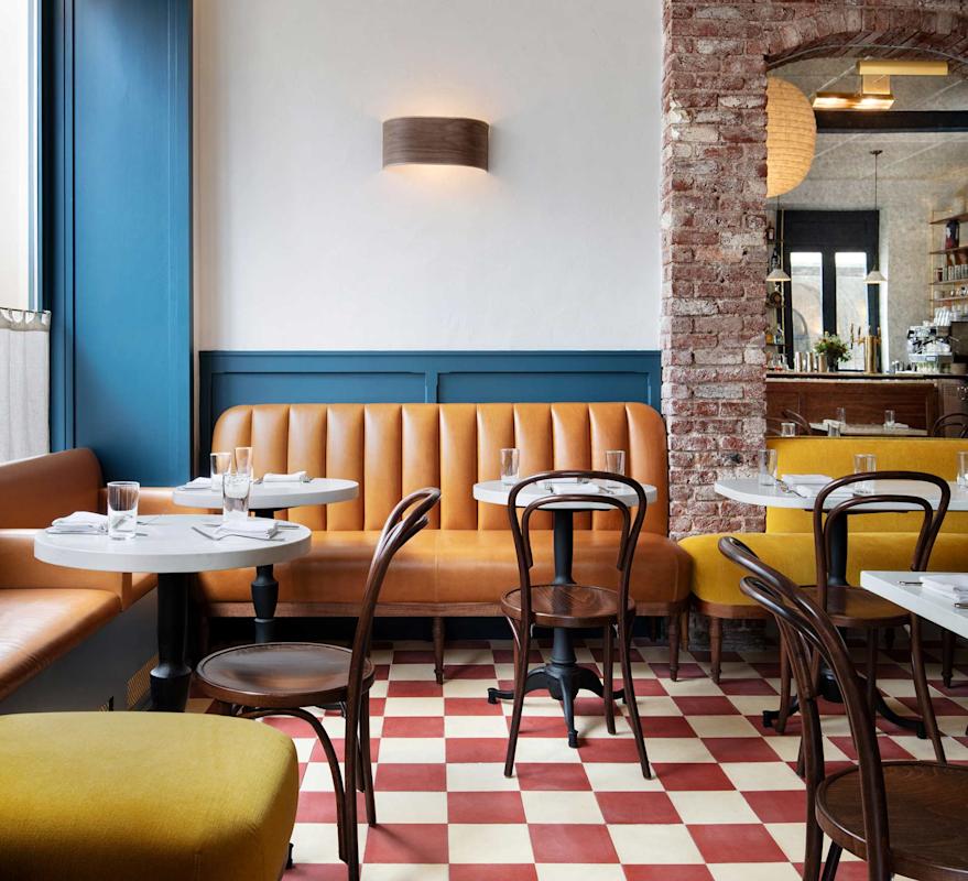 A cozy restaurant interior with tan leather banquette seating, round marble tables, wooden chairs, red and white checkered floor tiles, exposed brick wall, and a modern wall sconce.