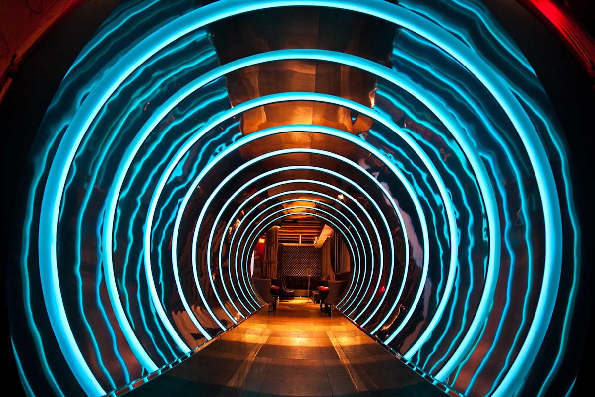 A futuristic hallway features concentric blue neon rings illuminating the tunnel, leading to a cozy seating area with chairs and warm lighting at the far end. The walls and floor have a metallic, modern appearance.