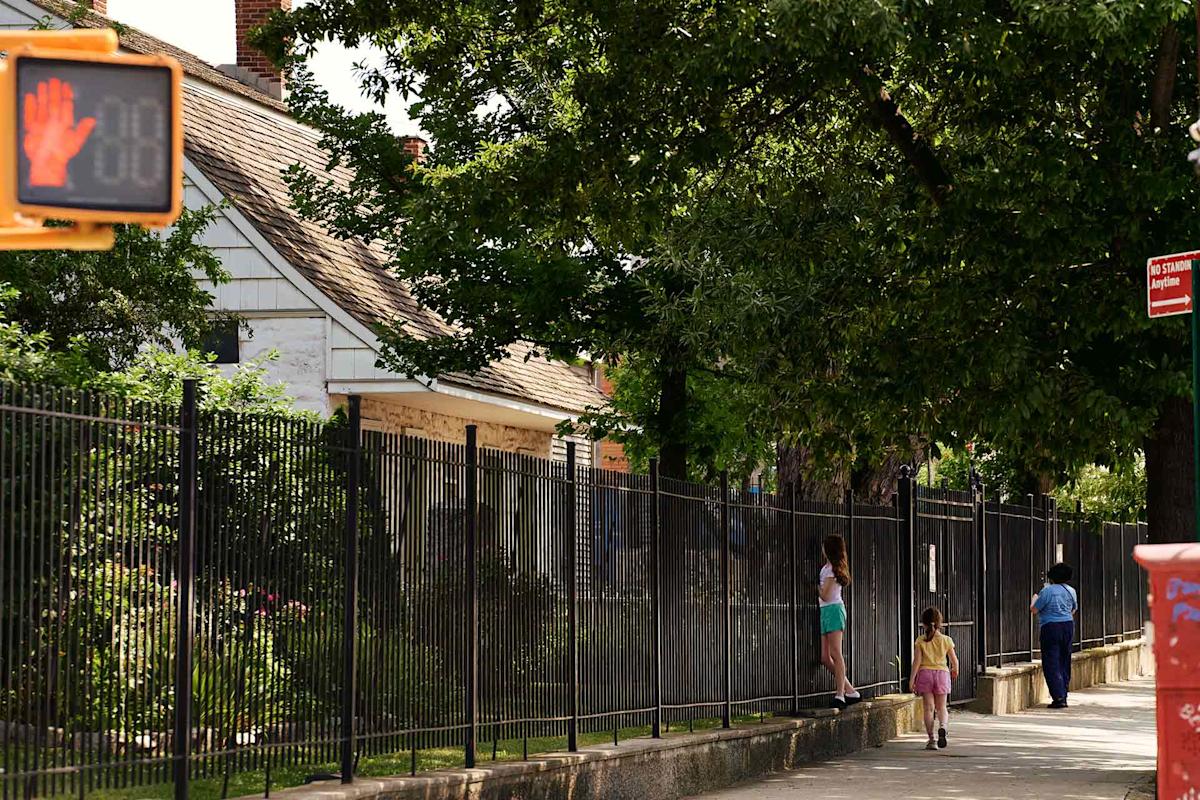 A sidewalk lined with a black fence beside houses, two children and an adult walking. A traffic signal shows a red hand and countdown. Trees provide shade over the scene on a sunny day.