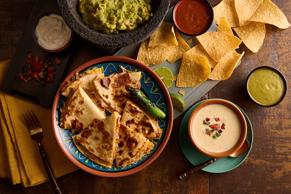 A colorful table spread with cheese quesadillas, tortilla chips, guacamole, salsa, queso dip, a grilled jalapeño, sour cream, and chopped tomatoes on a rustic brown surface.