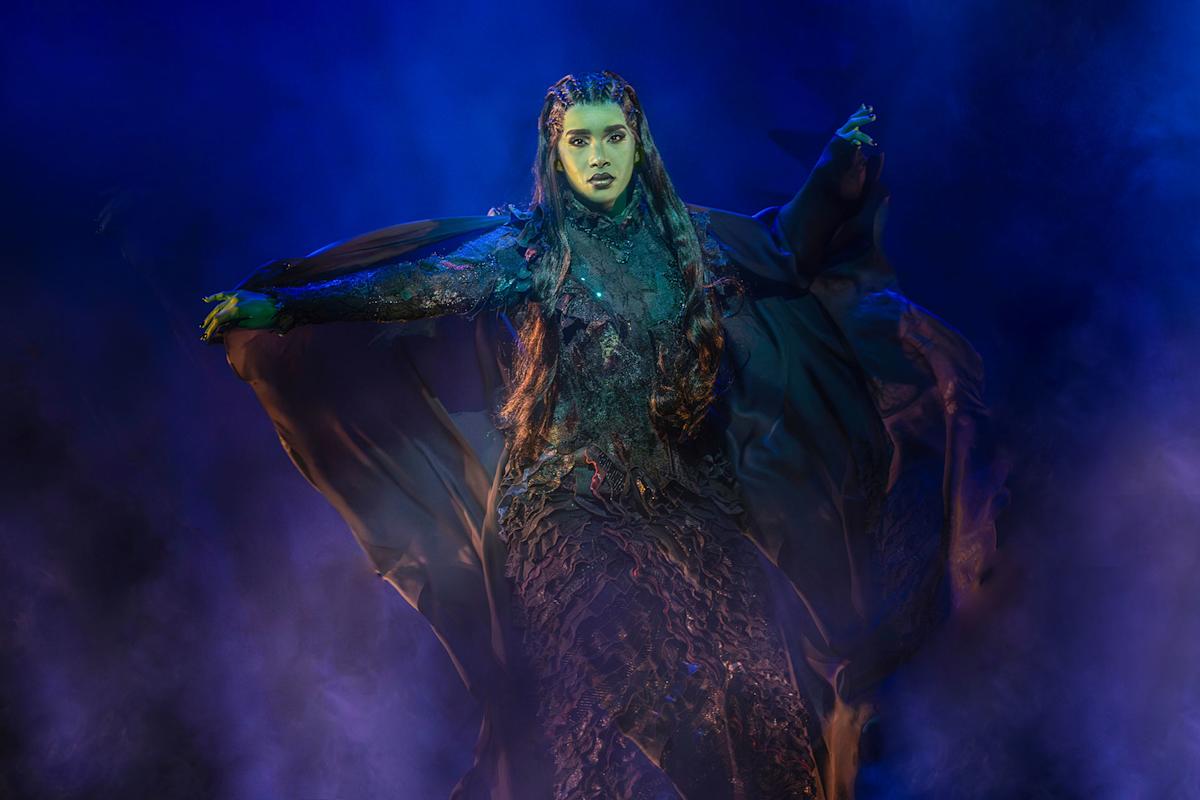A woman dressed as a witch in a dramatic, dark, textured gown stands with arms outstretched amid swirling blue and purple smoke on stage, her face painted green and expression intense.