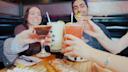 Four people sitting at a restaurant table clink their colorful cocktails together in a toast. The table has food, and everyone is smiling, creating a cheerful and lively atmosphere.
