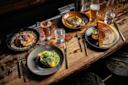 A rustic wooden table set with four gourmet dishes, two glasses of water, a glass of beer, and gold utensils. The dishes include pancakes, eggs on toast, a crepe with sauces, and a salad with greens.
