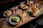 A rustic wooden table set with four gourmet dishes, two glasses of water, a glass of beer, and gold utensils. The dishes include pancakes, eggs on toast, a crepe with sauces, and a salad with greens.