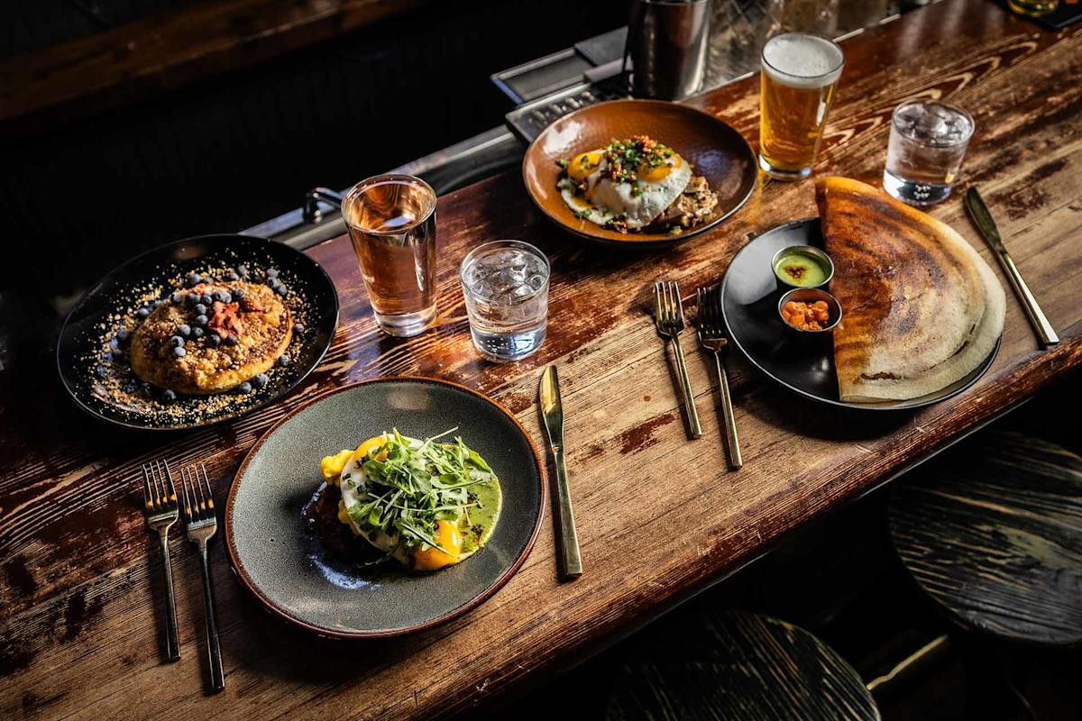 A rustic wooden table set with four gourmet dishes, two glasses of water, a glass of beer, and gold utensils. The dishes include pancakes, eggs on toast, a crepe with sauces, and a salad with greens.