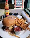 A plate with a cheeseburger, crispy French fries, and a cup of ketchup sits on a table next to a bottle of Louisiana hot sauce, a margarita, and sunglasses; a napkin is rolled beside the plate.