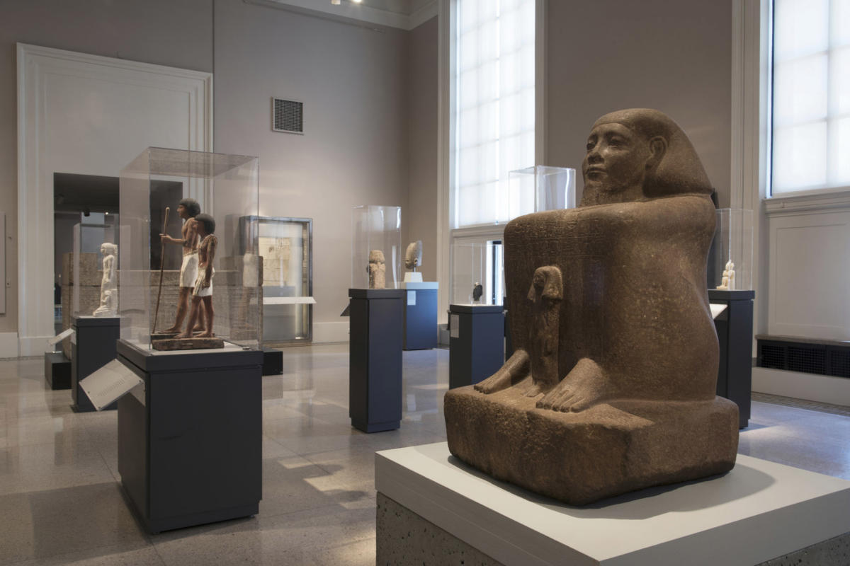 A museum gallery displaying various ancient Egyptian sculptures and artifacts, including a large stone statue of a seated figure in the foreground. The room is softly lit with natural light from large windows.