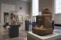 A museum gallery displaying various ancient Egyptian sculptures and artifacts, including a large stone statue of a seated figure in the foreground. The room is softly lit with natural light from large windows.