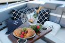 A table on a yacht displays croissants, strawberries, grapes, and apricots on a platter, with two empty champagne flutes and a bottle of champagne in an ice bucket. Nautical pillows and blankets decorate the seating area.
