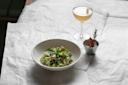 A bowl of fresh green salad with nuts sits on a white surface next to a cocktail in a stemmed glass and a small metal cup garnished with rosemary and dried fruit.