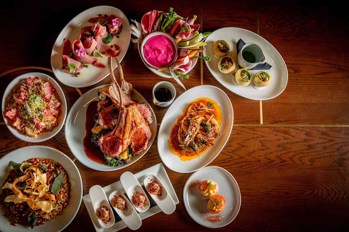 An overhead view of a table set with colorful dishes, including lamb chops, deviled eggs, pink dip, seafood, salads, and garnished plates, all arranged on white plates on a wooden surface.