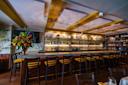 A modern bar with a long marble countertop, high wooden chairs, shelves stocked with bottles, warm lighting, and a large floral arrangement on the left side. Decorative pendant lights hang from the ceiling.