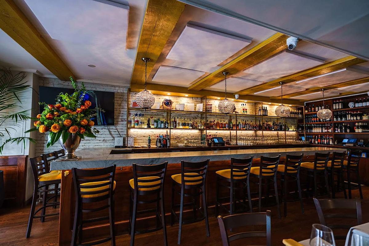 A modern bar with a long marble countertop, high wooden chairs, shelves stocked with bottles, warm lighting, and a large floral arrangement on the left side. Decorative pendant lights hang from the ceiling.