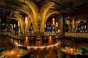 A modern, dimly-lit bar with intricate, towering light fixtures shaped like tree trunks. The bar is surrounded by green stools, and tables with green chairs and set dining ware. The ambiance is warm and elegant.