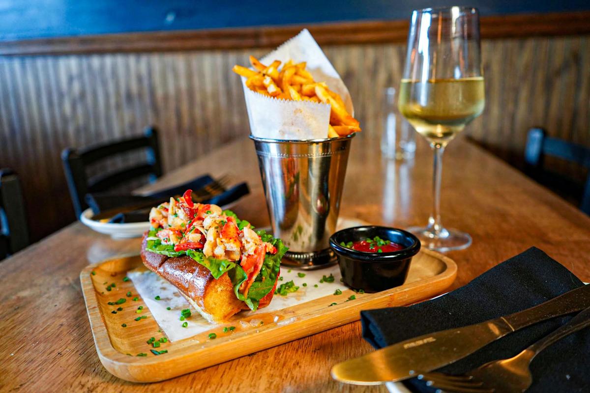 A lobster roll with lettuce on a wooden tray, served with fries in a metal cup, a small container of sauce, and a glass of white wine on a wooden table.