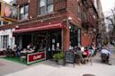 People dine outdoors at a sidewalk café called Claret on a city corner. The café has string lights, a red awning, and a chalkboard menu. A traffic signal and brick apartment buildings are visible in the background.