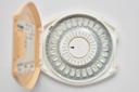 An open beige case containing a circular pack of birth control pills, labeled with the days of the week in a ring, with an arrow pointing to a specific day. The background is plain white.