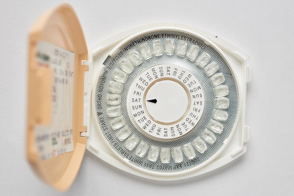 An open beige case containing a circular pack of birth control pills, labeled with the days of the week in a ring, with an arrow pointing to a specific day. The background is plain white.