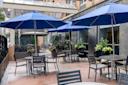 Outdoor dining area with several tables and chairs under blue umbrellas. Potted plants decorate the space, which is enclosed by a modern fence. A bicycle rack and building exterior are visible in the background.
