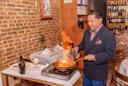 A well-dressed man flambés food in a pan at a restaurant table, producing a large flame. The setting features brick walls, a wine display, and elegantly set tables.