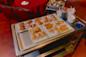 A metal cart holds plates of various dim sum items, including spring rolls, fried dumplings, sesame balls, and pastries, ready to be served in a restaurant with red chairs and tables in the background.