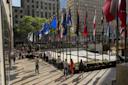 People walk along a plaza lined with many international flags at Rockefeller Center in New York City. Tall buildings and greenery are visible in the background under clear, sunny skies.