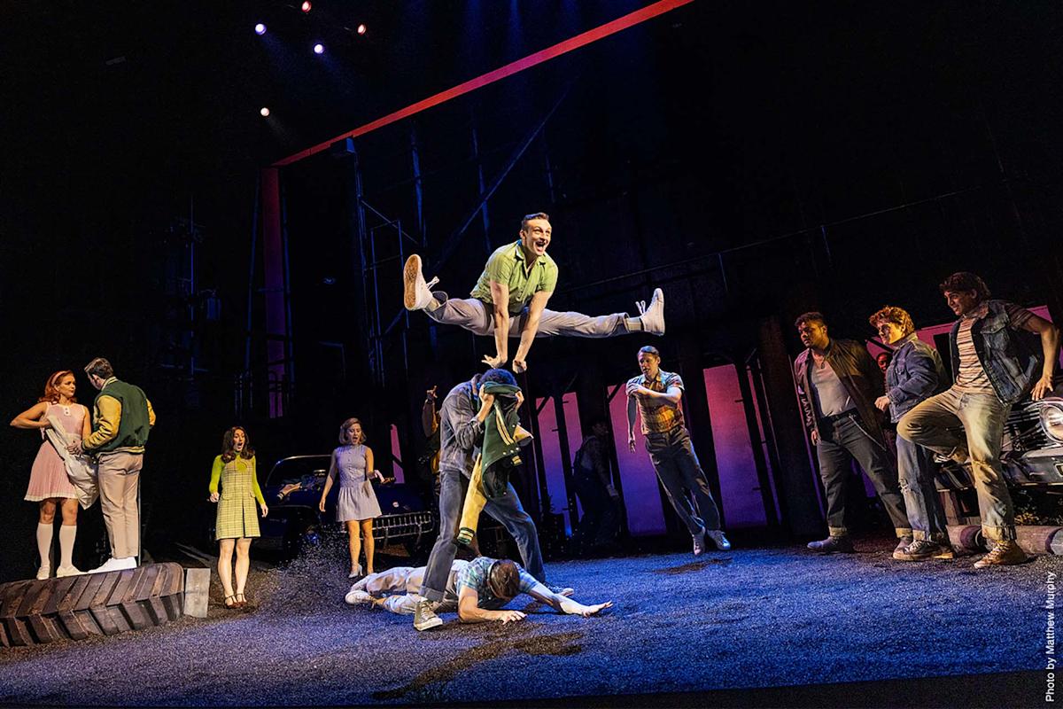 A lively stage scene shows a performer leaping over another cast member, with others watching. The set features cars, dramatic lighting, and characters in retro 1960s-style attire, evoking an energetic musical performance.