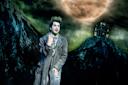 A person in tattered, gray clothing with green face paint and wild hair stands in front of a spooky, illustrated backdrop featuring a large full moon and a crooked house on a hill.