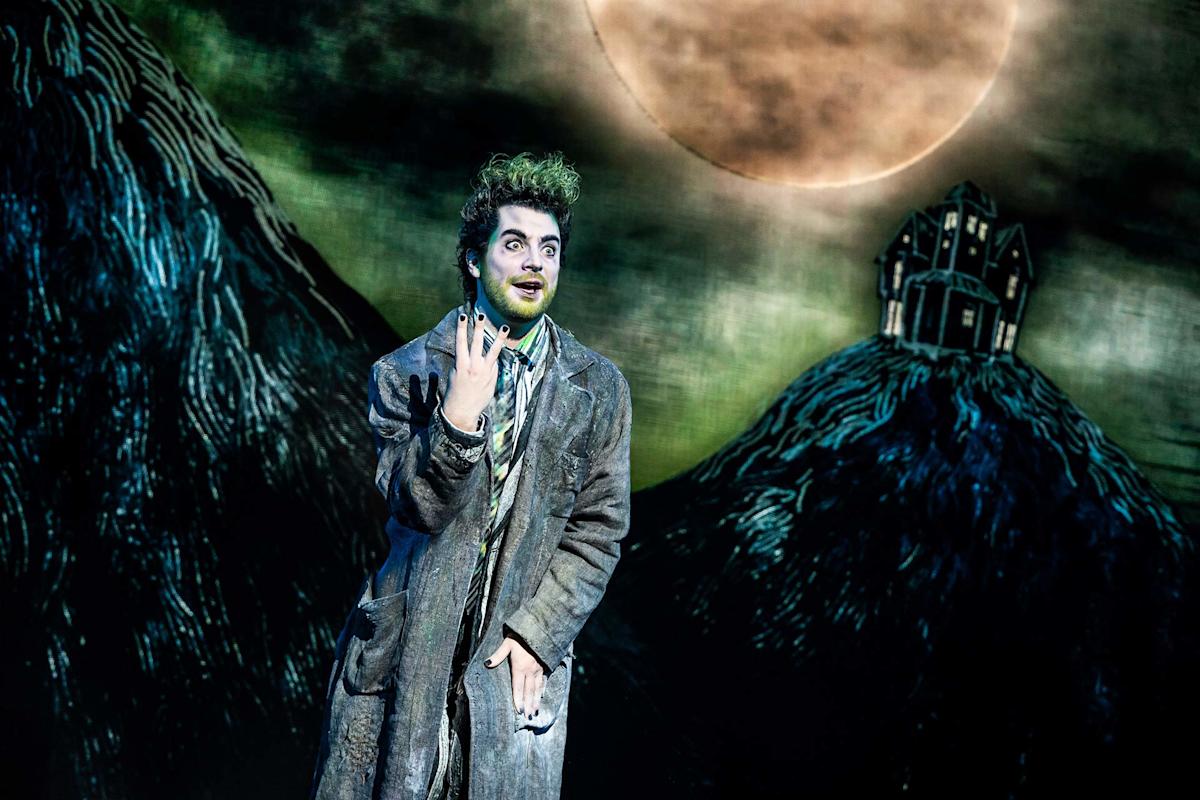 A person in tattered, gray clothing with green face paint and wild hair stands in front of a spooky, illustrated backdrop featuring a large full moon and a crooked house on a hill.