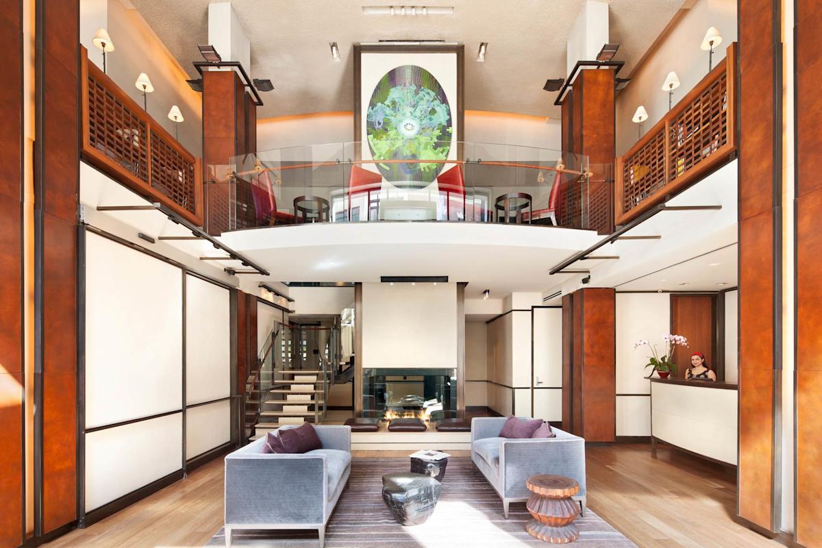 A modern hotel lobby with two gray sofas, decorative tables, a fireplace, and a mezzanine with glass railing. Warm wood accents and a large abstract artwork hang above the seating area.