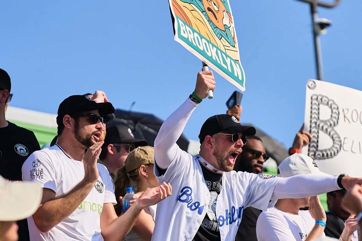 A group of enthusiastic fans in sunglasses cheer at an outdoor event under a clear sky. One man in front holds a cartoon sign reading "Brooklyn" while others clap and show excitement.