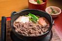 A hot pot dish featuring sliced beef, tofu, green onions, and tomato in a black pot. In the background, there's a red bowl with soup and a small dish with greens, all set on a wooden surface.