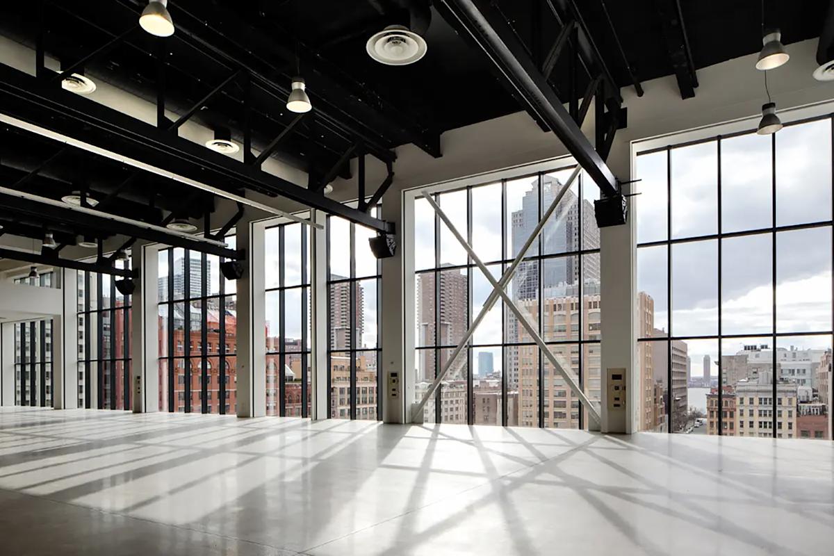 A spacious, empty loft with large floor-to-ceiling industrial windows, black steel beams, and city skyscrapers visible outside in daylight. Sunlight casts geometric shadows on the polished concrete floor.
