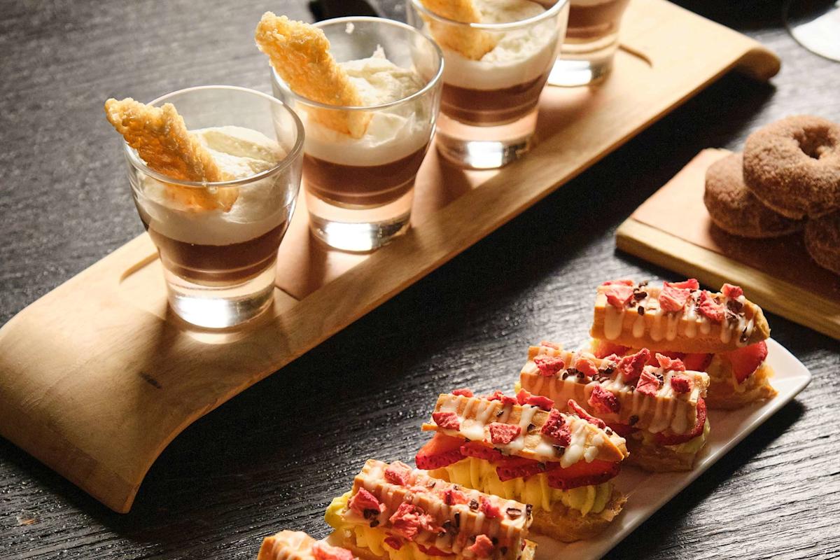 A wooden tray holds glasses filled with layered dessert and a biscuit stick, while a plate displays éclairs topped with cream and strawberries. In the background are small sugar-coated donuts.