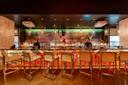 A modern, stylish bar with six high chairs, illuminated shelves of liquor bottles, two bartenders, and a large mural of a woman's face behind the bar. Warm lighting and marble decor create an upscale atmosphere.