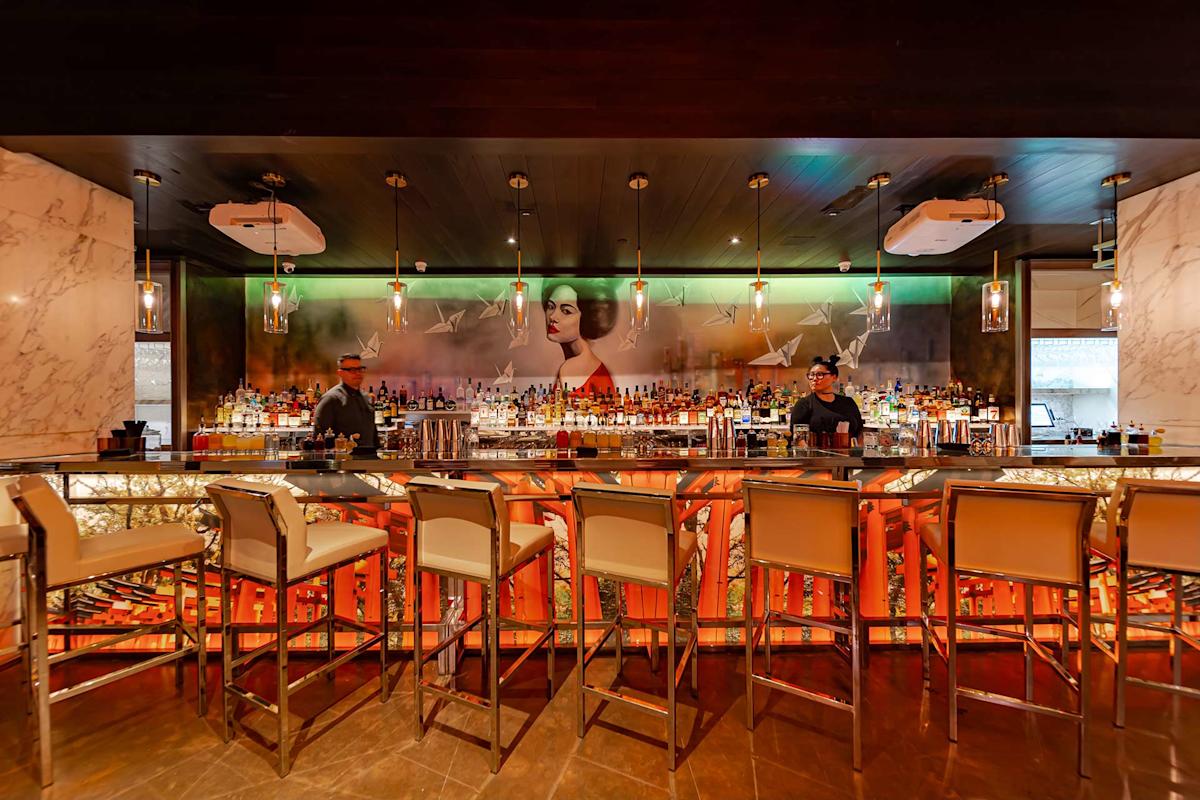 A modern, stylish bar with six high chairs, illuminated shelves of liquor bottles, two bartenders, and a large mural of a woman's face behind the bar. Warm lighting and marble decor create an upscale atmosphere.
