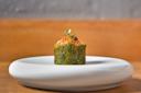 A single piece of sushi wrapped in seaweed, topped with orange fish roe, microgreens, and garnishes, is presented on a minimalist white plate with a blurred wooden and gray background.