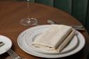 A neatly set dining table features a plate with a folded beige napkin labeled "Café Boulud," cutlery, and a wine glass on a wooden surface with a green cushioned background.